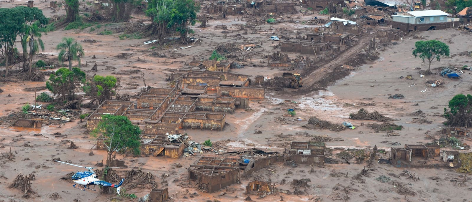 Ampla vista aérea do povoado destruído, com rejeitos cobrindo o solo entre as construções em ruínas.