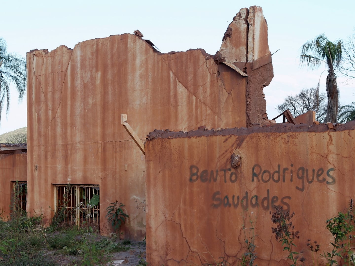 Um muro destruído e manchado de lama no povoado esvaziado de Bento Rodrigues, com as palavras "Bento Rodrigues Saudades" escritas à mão, e a escola do povoado destruída ao fundo.